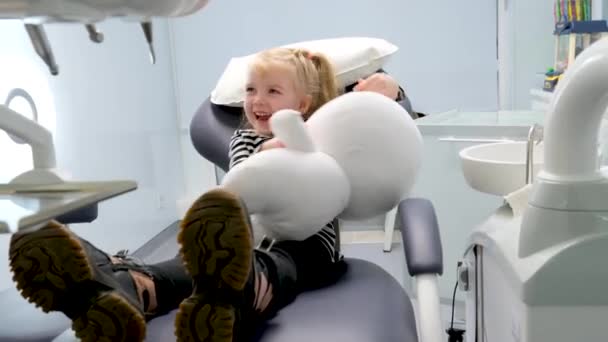 小さな女の子の歯科椅子を示す若い医師歯科医への最初の旅行幸せな笑顔楽しい会話ミルク歯治療看護師フェアヘア女の子の歯科事務所最新の技術 — ストック動画
