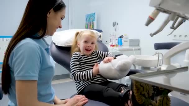 小さな女の子の歯科椅子を示す若い医師歯科医への最初の旅行幸せな笑顔楽しい会話ミルク歯治療看護師フェアヘア女の子の歯科事務所最新の技術 — ストック動画