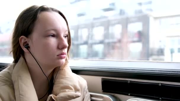 十代の女の子乗ります電車の中でバスヘッドフォンで耳の中の音楽に耳を傾ける秋の冬暖かいベージュのジャケット髪は下に縛られています本当の人座っている悲しい夢の下に隠された足待っています停止 — ストック動画