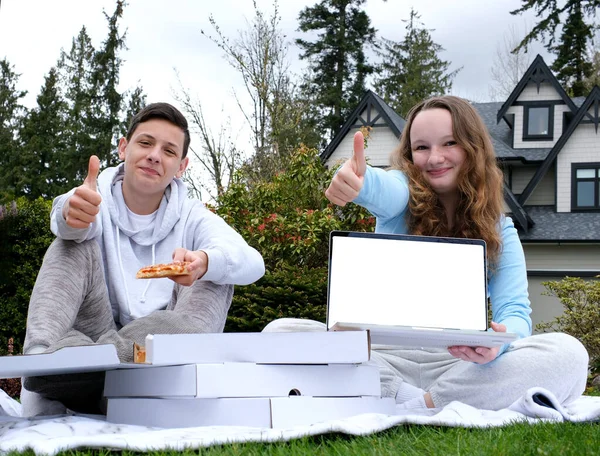teenagers are holding mockup laptop, cheerful girl and boy are holding mockup laptop. Blank white screen of modern notebook. I smile, I raise my finger up recommend site. Sitting on grass picnic pizza