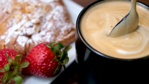 泡とおいしいカプチーノストロベリーとクロワッサンは粉砂糖を振りかけた素敵なフランスの朝食スプーンと攪拌コーヒー滴がスローモーションブラックカップホワイトスクエアプレートを落下 — ストック動画