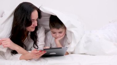 Anne ve oğlu battaniyenin altında 4-6 yaşlarındaki tablet çocuk üzerinde oynuyorlar siyah saçlı güzel bir kadın beyaz çarşaf altında yatıyor kahkaha atarak gülüyor internet üzerinden eğitim aktiviteleri öğreniyor