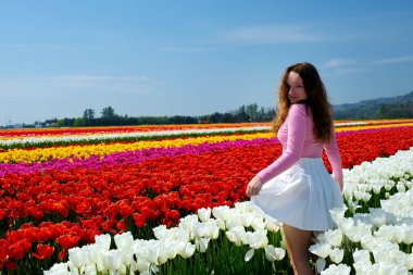 Kız tarlada dönüyor laleler rüzgâr eteğini kaldırıyor arka planda çiçekleri tutuyor neşe içinde gülüyor gençlik temizliği saç maviliği berrak gökyüzü temizliği temiz bekaret