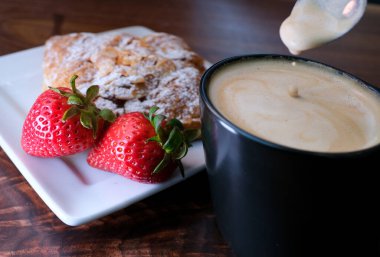 Lezzetli kapuçino köpüklü çilek ve üzerine pudra şekeri serpiştirilmiş kruvasan harika Fransız kahvaltısı kaşığı ve karıştırılmış kahve damlaları yavaş çekimde siyah fincanın beyaz kare tabağına dökülüyor.
