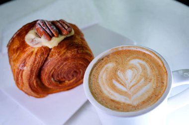 Lokantada nefis bir tatlı, beyaz tabakta yuvarlak kruvasan, cevizli ve kremalı pasta, vanilyalı kouign Amann yuvarlak kruvasan, New York Roll, tarçınlı tarçınlı latte.