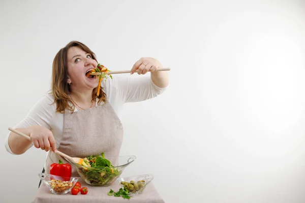 funny cheerful woman eating salad on white background she opens mouth wide bulges eyes large wooden spoon stuffs portion of food on table ingredients space for text weight loss ad