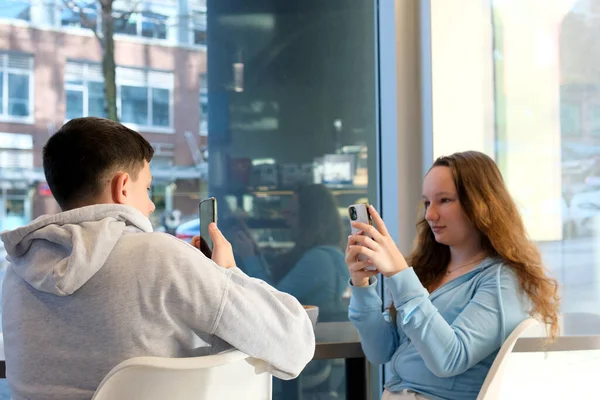 男の子と女の子がそれぞれのスマートフォンで窓のそばのテーブルに座っている携帯電話の十代の若者の生活は Fiがあるときにソーシャルネットワークでインターネット上で時間を過ごす灰色の青い白い色 ストックフォト