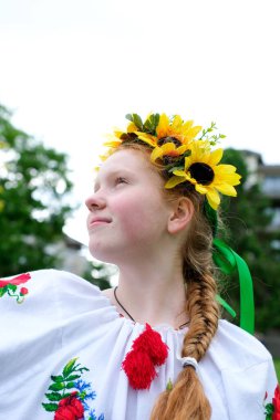 Parlak kızıl saçlı, güneşli bir kız, yeşil ağaçların arkasında duruyor. Üzerinde ayçiçeği çelengi işlenmiş Ukraynalı kadın barış için Ukraynalı kadın. Savaş zaferi Ukrayna ulusal vatanseverliği değil.