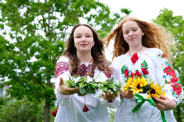 2人の美しい若い女の子が刺繍のシャツを着て笑って笑顔の花輪を水の上にカメラが私たちに花輪を伸ばしに直面して私たちに異なるひまわりの女の子が持っている赤い明るい日当たりの良い髪 ストック画像