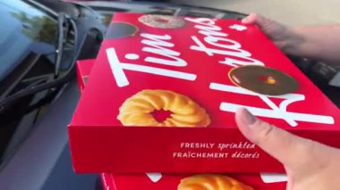Kontrastlı ışıkta Tim Hortons çöreklerinden oluşan geniş, açık bir kutu. Bir kadın hediye olarak iki kutu lezzetli tatlı taşır, bir ikram, Kanada Vancouver 2023