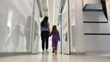 Kadın ve kız koridor boyunca yürür birbirlerine bakarlar büyük beyaz koridor şeffaf beyaz kapı yansıması hastane bakım konsepti çocuklara yardımcı olur. 