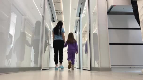 女性と女の子が廊下に沿って歩くお互いを見て大きな白いホール透明なガラスの白いドアの反射病院のケアコンセプトは子供たちを助ける — ストック動画