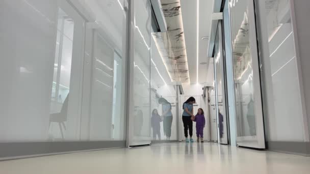 女人和女孩走在走廊上互相望着大白厅透明的玻璃门反射医院护理理念帮助孩子们 — 图库视频影像