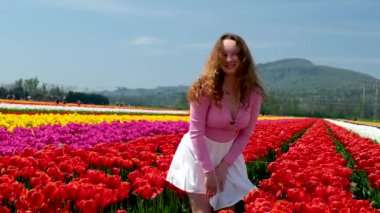 Beyaz tenis eteği içinde kırmızı laleler içinde kıvrılan genç bir kadın. Güzel pembe bluz. Uzun saçlı, kahverengi saçlı kadın el ele tutuşuyor.