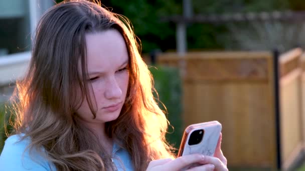 日落时 一个女孩用一个面容严肃的青少年的电话站在家里 阅读社交网络的信息通知 寻找地理定位许多技术和小工具的能力 — 图库视频影像