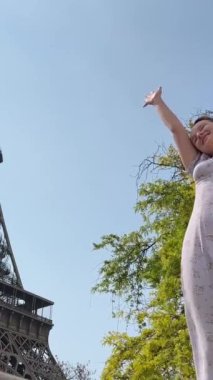 a beautiful teenage girl near the Eiffel Tower and a green tree and waves her hands with joy, she seems to be dancing and having fun there is a place for text about travel joy partying.