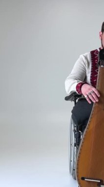 Elinde kobza enstrümanı olan yakışıklı genç adam tekerlekli sandalyede beyaz arka planda oturuyor bacakları olmayan Ukrayna nakışlı gömlek giymiş savaş gazisi. 