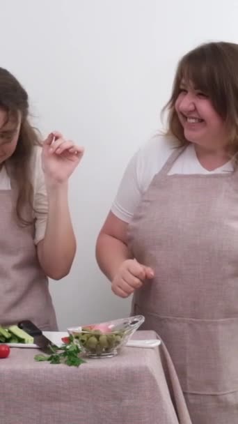 おいしいと健康的な食品を準備しながら2人の女性は踊ります手でベジタリアン食品野菜サラダ木製のスプーン同じキッチンエプロン白い背景喜び家族 — ストック動画