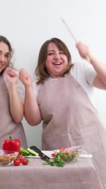 2 women dancing while preparing tasty and healthy food vegetarian food vegetable salad in hands wooden spoons identical kitchen aprons white background joy fun family Cozy weekend mom and daughter