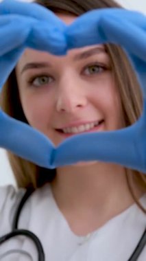 Doctor hands in gloves in the heart shape against the background of his body face and medical gown. closeup. Female hand in blue medical gloves show heart sign. High quality photo