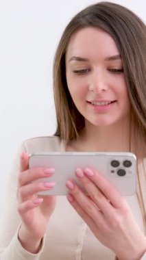 Avrupalı uzun saçlı, kahverengi saçlı güzel bir kız beyaz arka planda akıllı telefondan alışveriş yapıyor. Yüksek kalite 4k görüntü