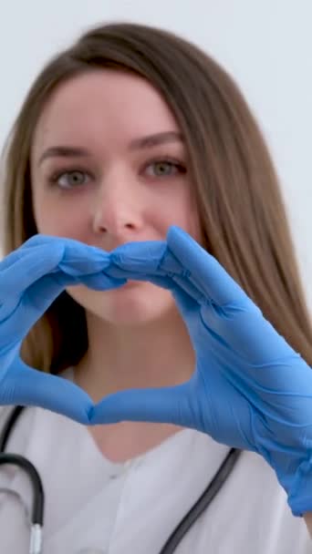 笑年轻的女医生心脏病医生身穿白色的医疗外套和听诊器显示手的心脏形状看着相机 心脏科保健 爱情和医药慈善概念 — 图库视频影像