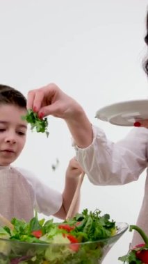 Güzel ev hanımı, küçük oğlunun taze marul yapraklarını parçalayıp açık modern mutfakta salata pişirmesine bakıyor.