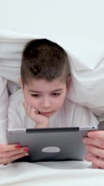 people, family and technology concept mother father and little boy with tablet pc computer in bed at home or hotel room. Happy family spend time together lying under white blanket on bed