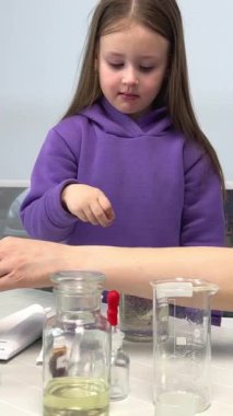 Chemistry laboratory girl in lilac tracksuit throws a pill into a glass bottle with experience for bubbles and looks with expectation