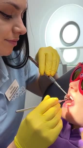 一个无法辨认的儿科牙医的手打电话给坐在医院椅子上的一个笑容可亲的可爱女孩 牙科医生 坐在牙医办公室黄色手套里的小女孩 — 图库视频影像
