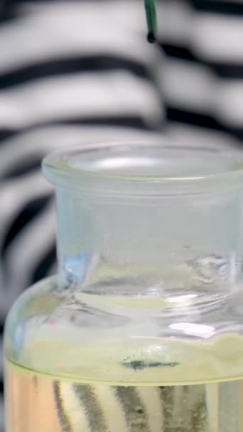 黄色の液体の青い緑色の滴を持つガラス瓶のクローズアップは縞模様のブラウスの背景に滴下しています — ストック動画