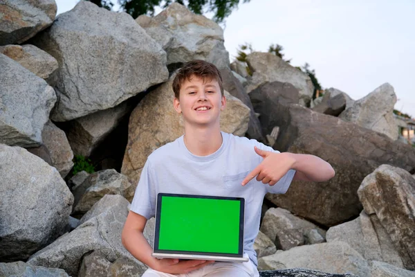 十代の男の子とノートパソコンで彼の手の石の山の上に座っている緑の画面ポイント指スペーステキスト幸せな笑顔グレーのTシャツ白のショートパンツは 広告楽しい喜びインターネットを提供します — ストック写真