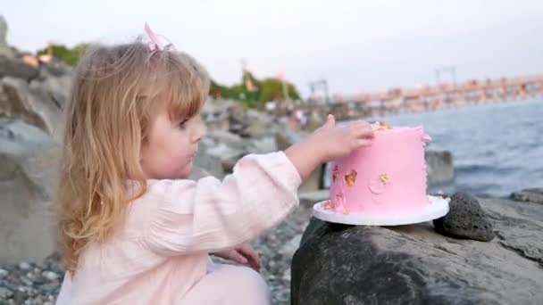 3岁的小女孩在海滩上吃着蛋糕过生日时吃着粉色的甜点 手里拿着三根蜡烛舔着的手指坐在石头上 靠近大海 — 图库视频影像