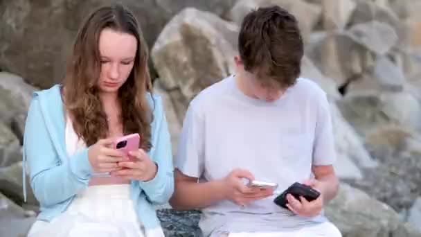 青少年们坐在石头上 男孩和女孩手里拿着手机 他们在自然界中使用依赖社交网络的小工具 — 图库视频影像