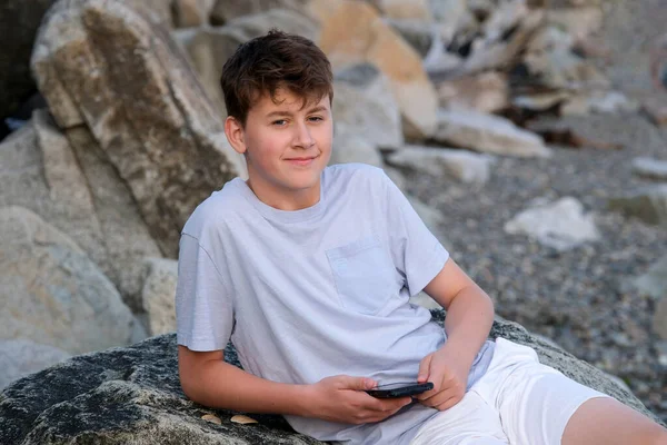 十代の男の子は石の海岸にビーチに横たわっていますビッグストーン晴れた日の海の残りのリラクゼーション夢の手下に頭のスニーカーを置きますショートパンツとTシャツ若い男性 — ストック写真