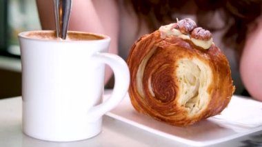 Kadın ısırılmış çöreği tutar, kafenin camından bakar, yansıtır. Ziyaretçi yüzü karede değil, akıllı telefonu sağ elinde tutuyor, mesaj yaz. Masanın üzerinde bir fincan kahve. Yüksek kalite