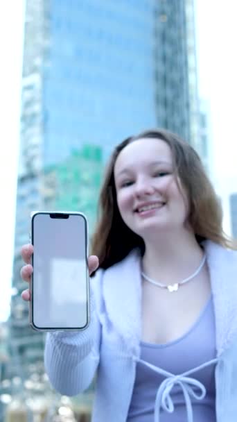 在摩天大楼的背景下 城市里的一位年轻女子用电话现场直播 看到一部手持白色摄像机的手机时 笑着把焦点放在手机的旁边 — 图库视频影像