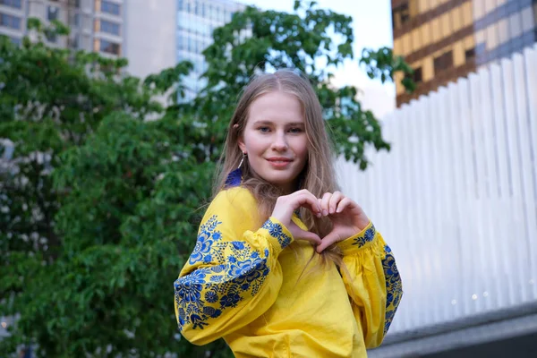 stock image cute beautiful blonde girl young woman looks into frame and makes a heart with hands. joyful in a yellow shirt on a white-green background street love peace in Ukraine joy calmness