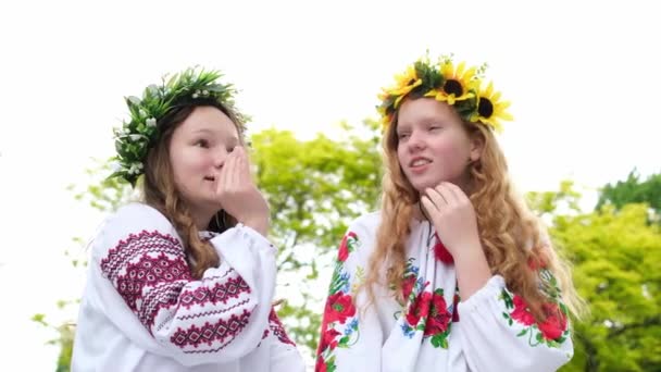 花輪を持つウクライナの女の子は 伝統的なウクライナの服に座っていると喜び 互いに耳の中に噂話をささやく興味深いことに聞く黒とブロンドの髪のガールフレンド民族衣装 — ストック動画