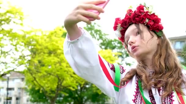 伝統的な民俗画像のウクライナの女性は 自撮りを取って電話を見ています 10代の女の子の多くの白い花の美しさと鏡の美しさの美しい花輪で見るウクライナ戦争の喜びの性質 — ストック動画