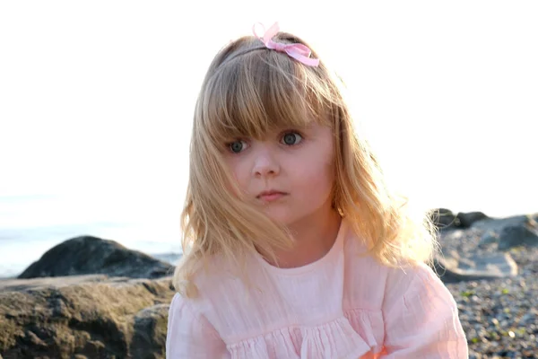 一个穿着粉红细腻衣服的小女孩坐在石头上 凝视着远处金色卷发孤寂的大海落日 — 图库照片