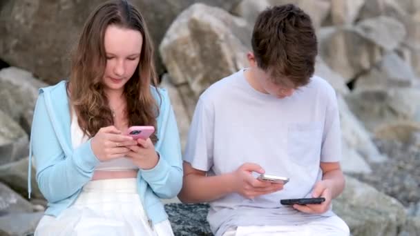 男の子の女の子のティーンエイジャーは 明るい服退屈な日付通信のダウンロードゲームは一緒に過ごす時間を通信再生します 青年期の電話中毒 — ストック動画