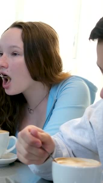 用勺子喝咖啡 一杯热饮男孩和女孩坐在咖啡店里 嘴里塞满了浓密的卡布奇诺咖啡泡沫 — 图库视频影像