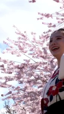 Güzel Asyalı kadın gülüyor rüzgarda saçlarını savuruyor kiraz çiçeklerine mavi gökyüzü beyaz bulutlar işlemeli Japon ulusal elbisesi 