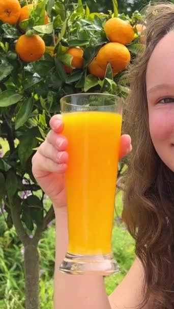 タンジェリンの木を背景にオレンジジュースを飲む女の子はオレンジジュースを飲むタンジェリンマンゴー彼女は貪欲に飲むと本当にどこでもおいしいジュースが好き緑と夏 — ストック動画