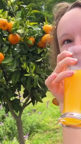 オレンジジュースのグラスを手にオレンジの木を背景に10代の少女がフレームにウィンクして顔を作る 高品質の写真 — ストック動画