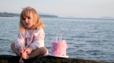 Pembe elbiseli küçük bir kız, doğum gününde lezzetli bir pembe pasta çiğnemeye bakıyor. 3 yıl deniz kıyısı güzelliği. Sevinç dolu aile kutlaması.,