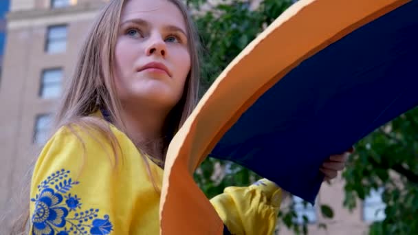 一个穿着绣花黄色衬衫 蓝色绣花的美丽的乌克兰女人举着乌克兰国旗 在房子的背景上 一棵树梦想着胜利的保护 请求帮助悲伤的眼睛 金黄的头发 — 图库视频影像