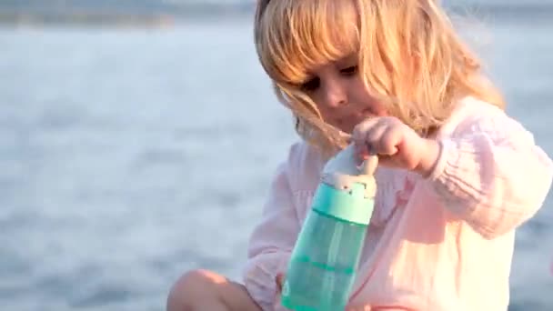 小女孩金发碧眼 穿着粉红的衣服 喝着蓝色瓶子里的水 营养正常 与大海的背景健康相符 — 图库视频影像