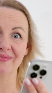 dikey video. Kadın beyaz arka planda mutlu bir şekilde gülümsüyor. Elleriyle telefonu tutuyor, alışveriş siparişi veriyor arkadaşlarıyla sohbet ediyor. Eğlenceli sohbetler.